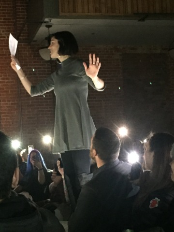Dessa reading a poem slightly blurry photo of a woman standing on a chair among seated people, reading
