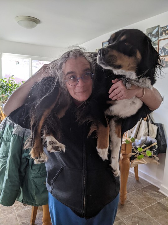 a gray-haired woman holding a black white and tan dog across her shoulders